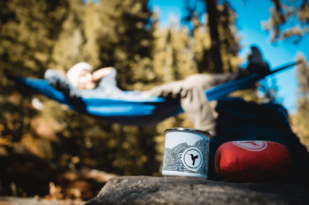 A person relaxes in a blue Hummingbird Hammock in a forest setting, with a focus on a Topo Enamel Mug and a red sleeping bag in the foreground.