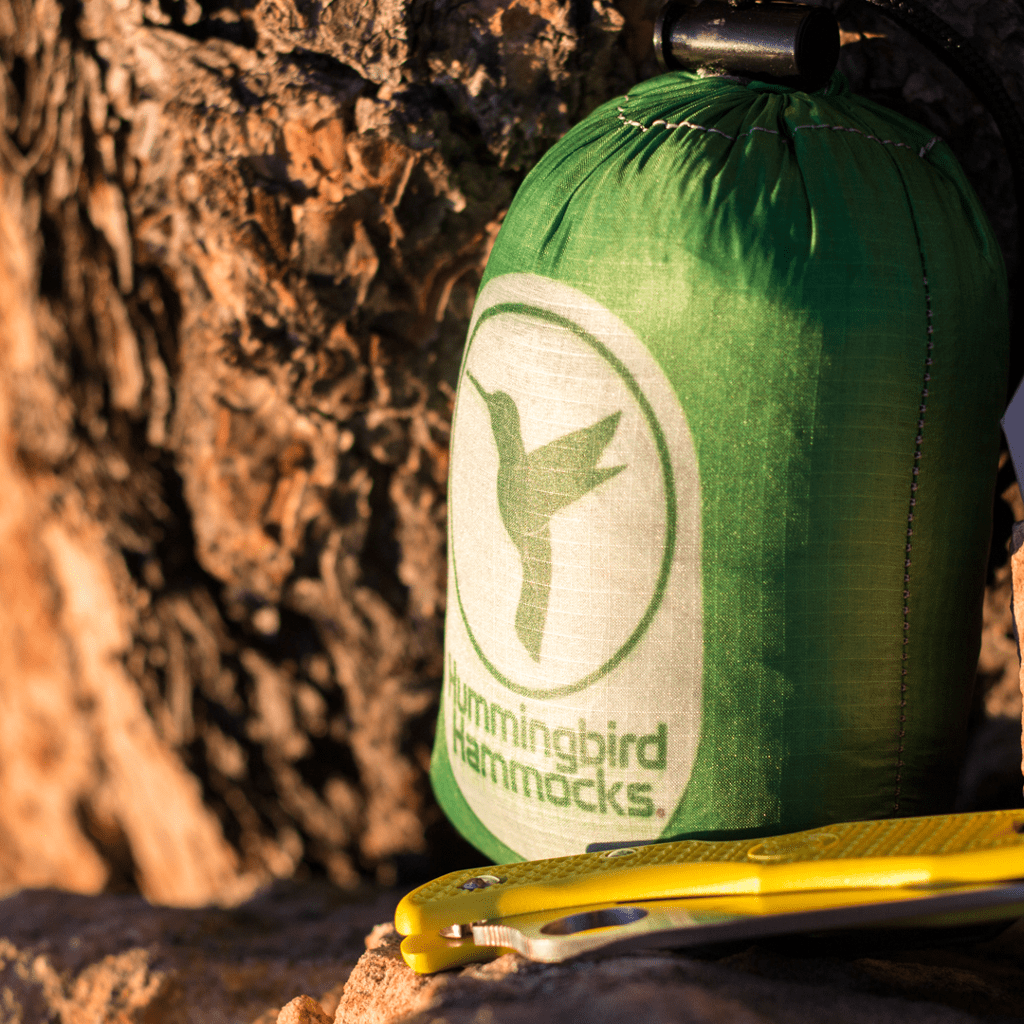 Green Ultralight Single Hammock by Hummingbird Hammocks packed in a bag, standing against a textured tree trunk, with a yellow knife beside it.