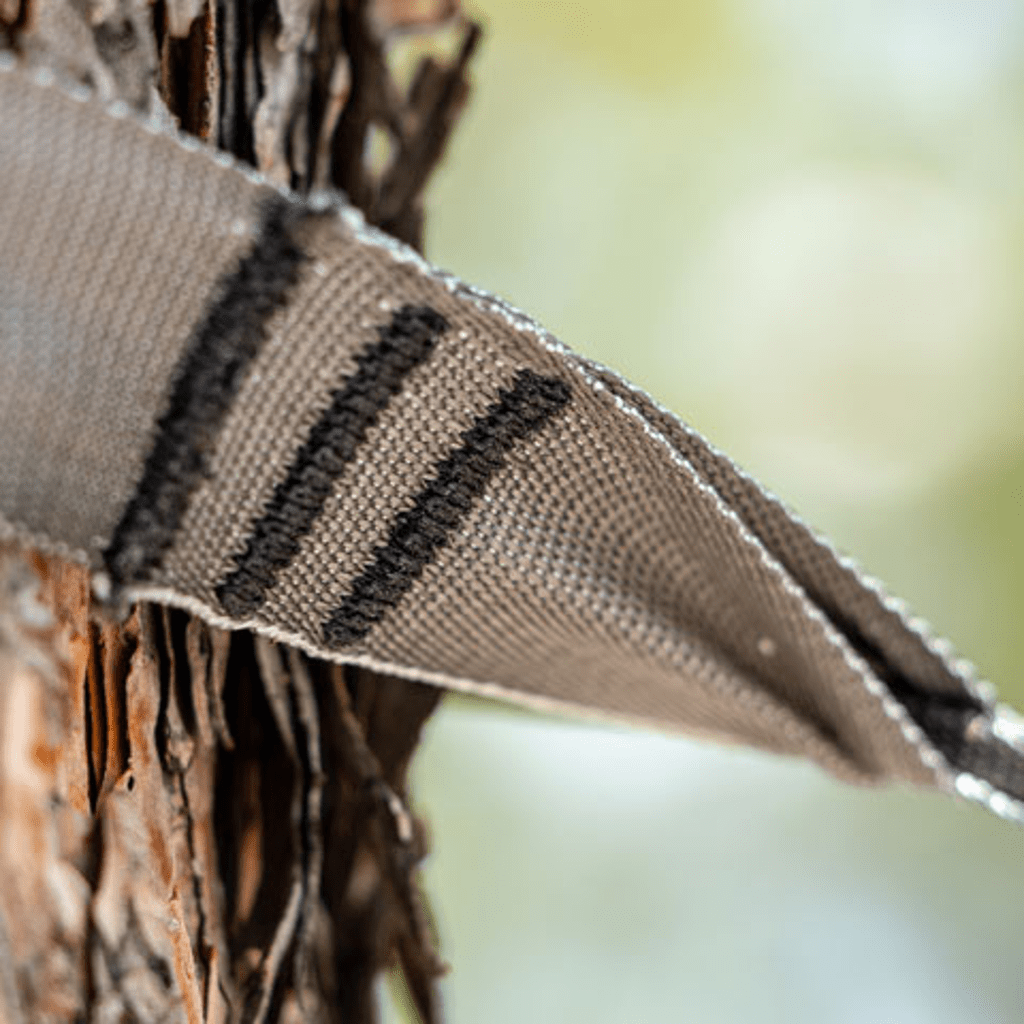 Close-up of beige and black striped Hummingbird Hammocks Tree Straps wrapped around a textured tree bark, with a blurred green background.